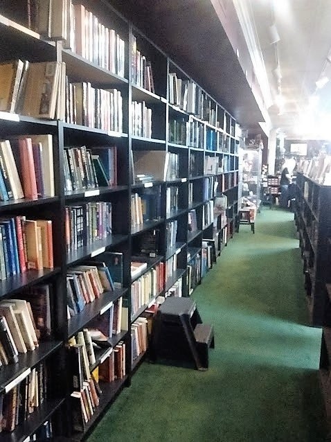 Long aisle of tall wooden bookshelves packed with books along a green carpet in a bookstore.