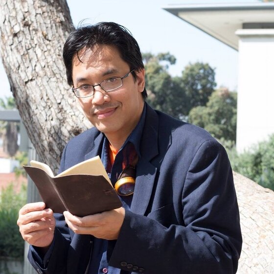 Bryan Thao Worra smiling and holding an open notebook outdoors under a tree