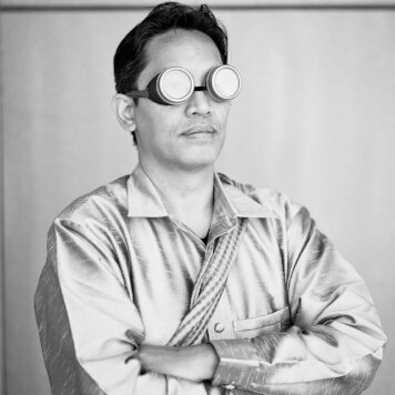 Black and white portrait of Bryan Thao Worra wearing round goggles and a silk shirt, arms crossed