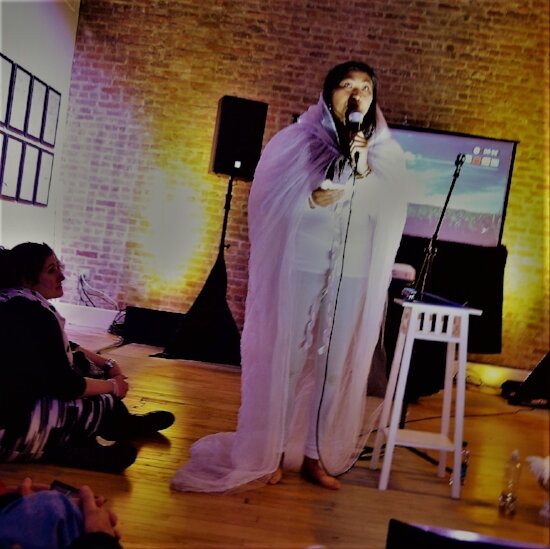Saymoukda Vongsay draped in white, reading into a microphone in a brick-walled performance space