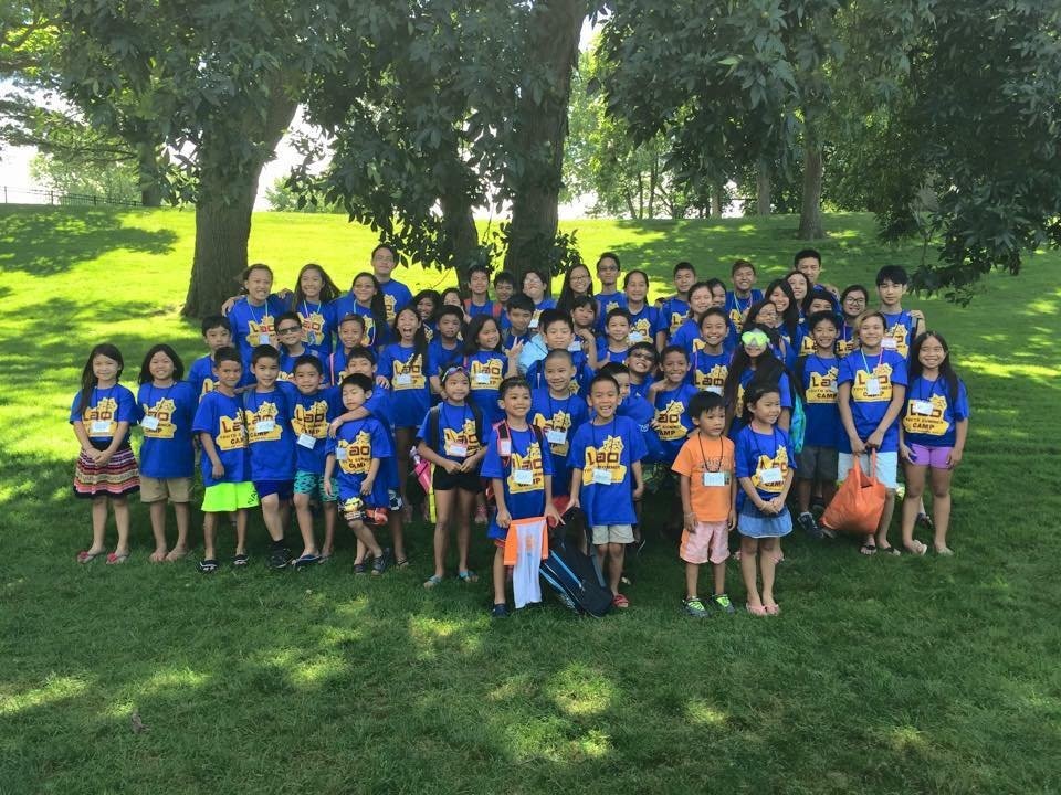 Group photo of children in matching blue Lao camp T-shirts on a grassy, tree-shaded lawn