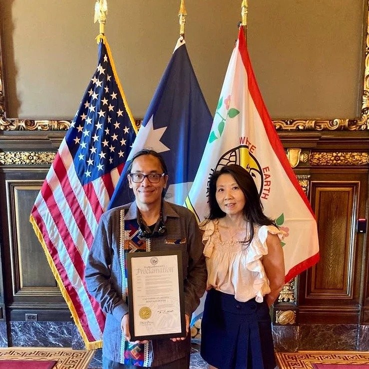 Two people holding the Lao American Artists Heritage Month proclamation in front of US and Minnesota flags