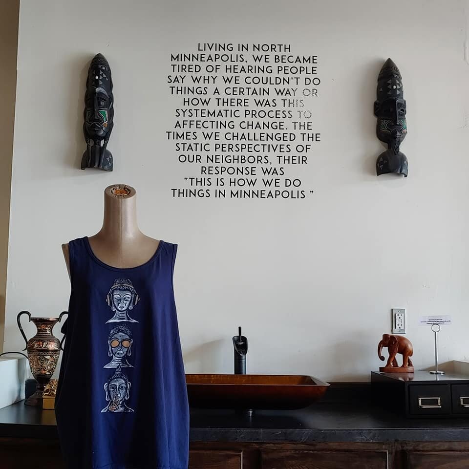 Gallery display with a tank top printed with three stylized Buddha heads, carved masks, and a wall quote about changing how things are done in North Minneapolis