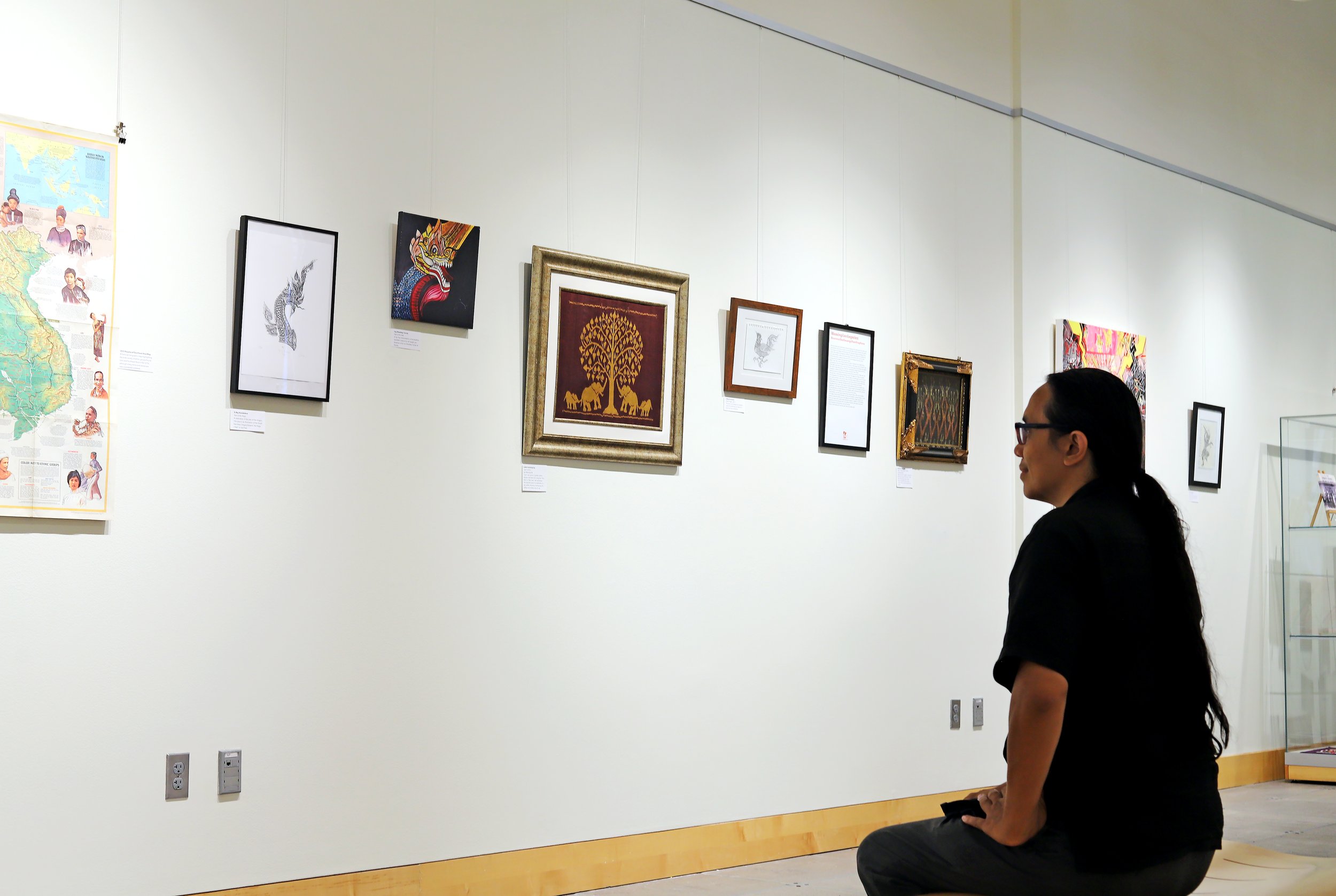 Visitor with long hair seated in a gallery, viewing a wall of Lao American artworks including a map and a gold tree textile