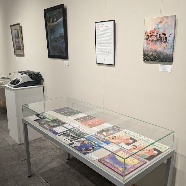 Laomerica 50 exhibit with a glass case of Lao American books, a vintage typewriter, and paintings on the walls