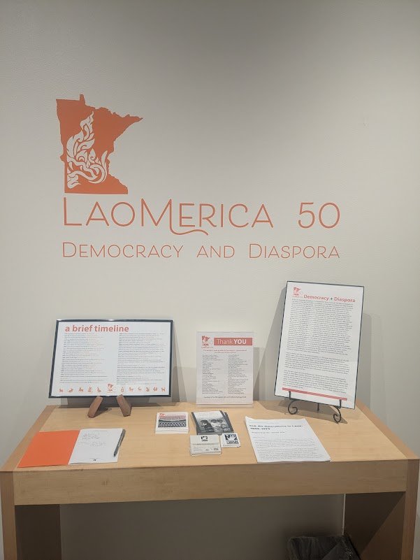 Laomerica 50: Democracy and Diaspora wall title above a table with a timeline, thank-you list, and handouts