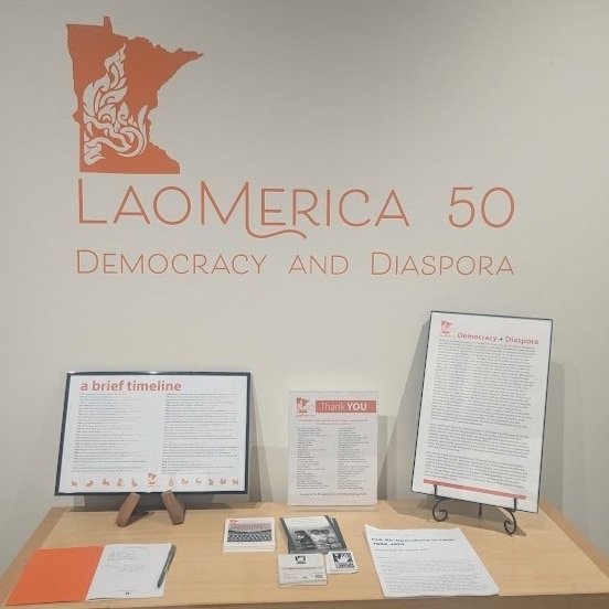 Laomerica 50: Democracy and Diaspora wall title above a table with a timeline, thank-you list, and handouts