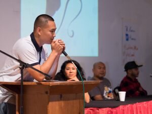Man speaking at a podium with panelists seated behind him at the Lao American Writers Summit