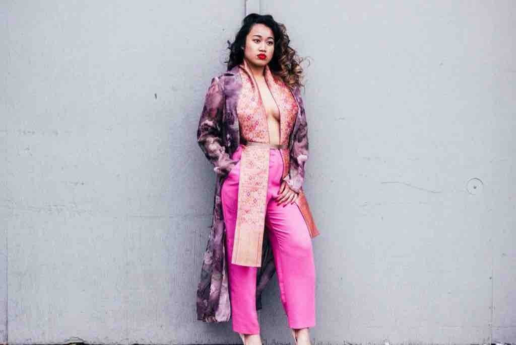 Lina V posing against a gray wall in a floral coat, pink trousers, and a patterned Lao textile sash