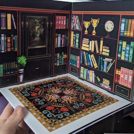 Hand holding open a paper diorama of a library room with colorful bookshelves, trophies, a globe, and a patterned rug.