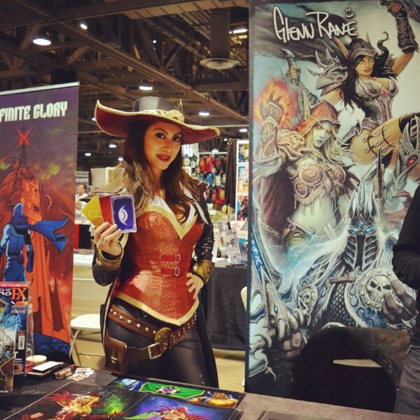 Woman in a red cowboy-style costume holding playing cards at a game booth beside a fantasy art banner