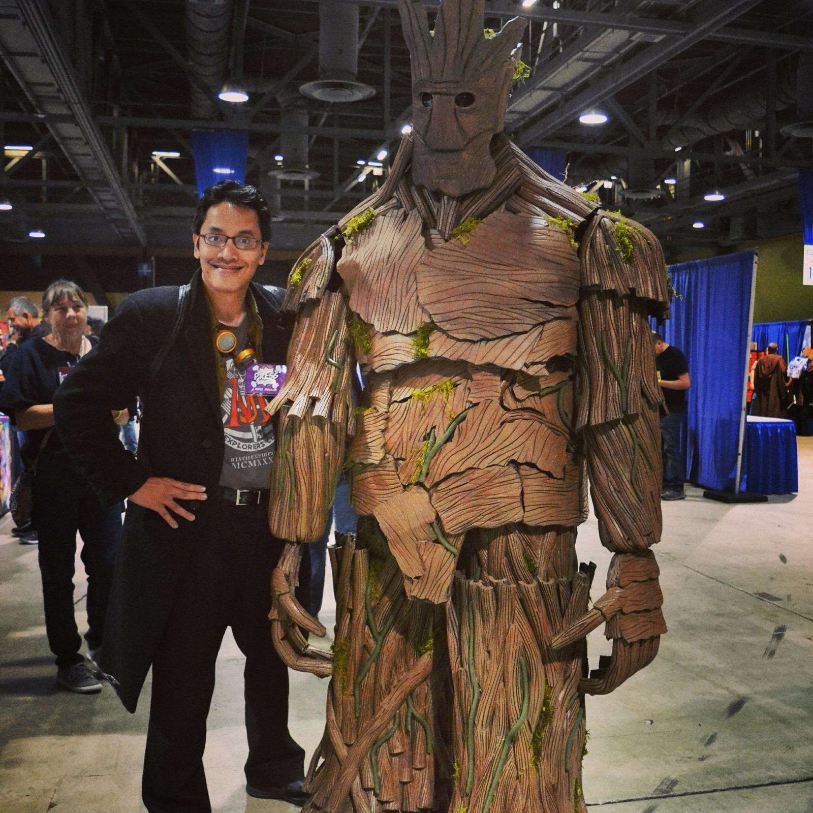 Smiling man in glasses posing beside a towering Groot costume on the expo floor