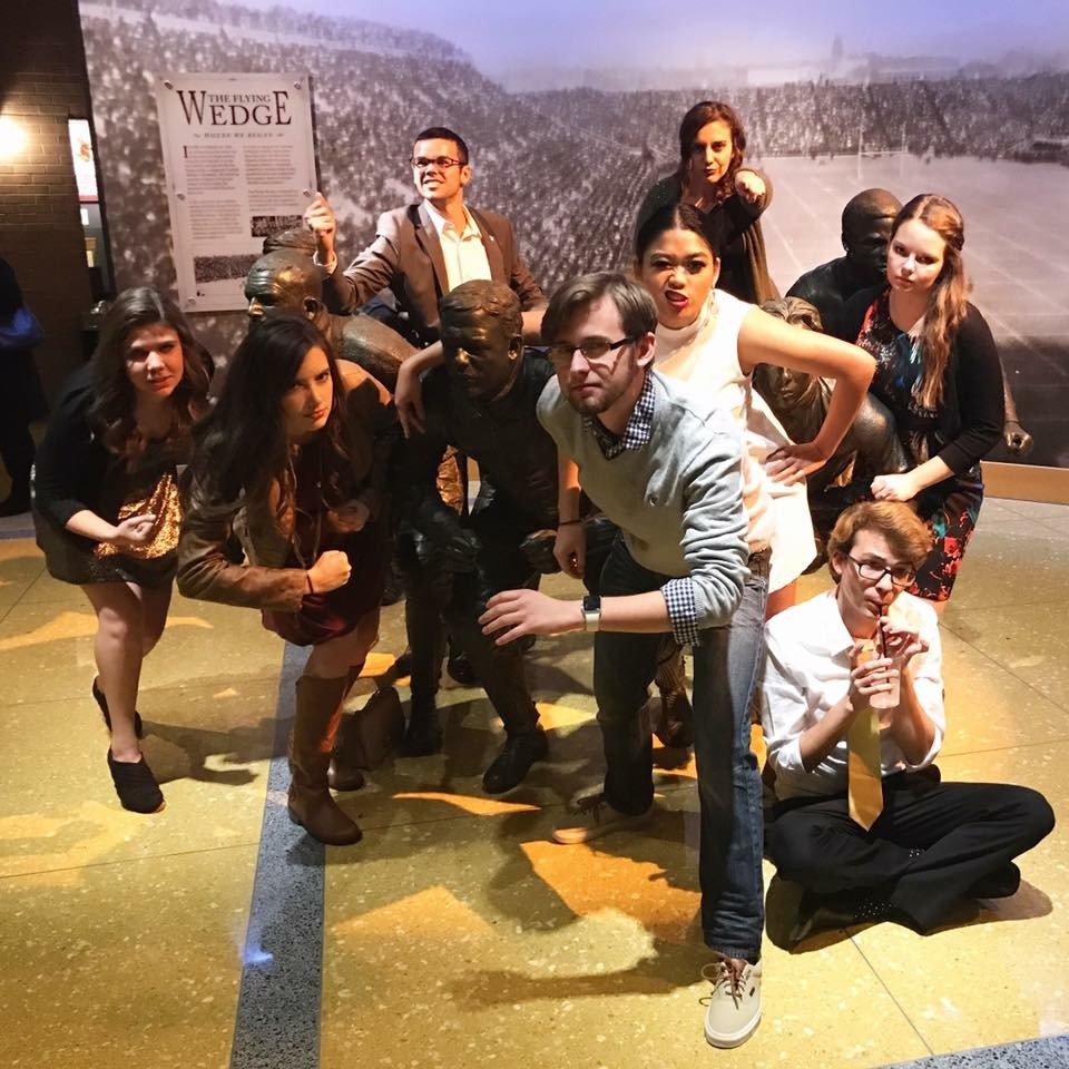 Group of young people striking playful football poses alongside bronze statues of football players in a museum.