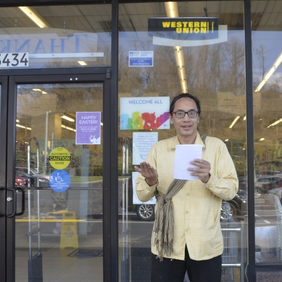 Bryan Thao Worra reading a poem from a sheet of paper outside a storefront