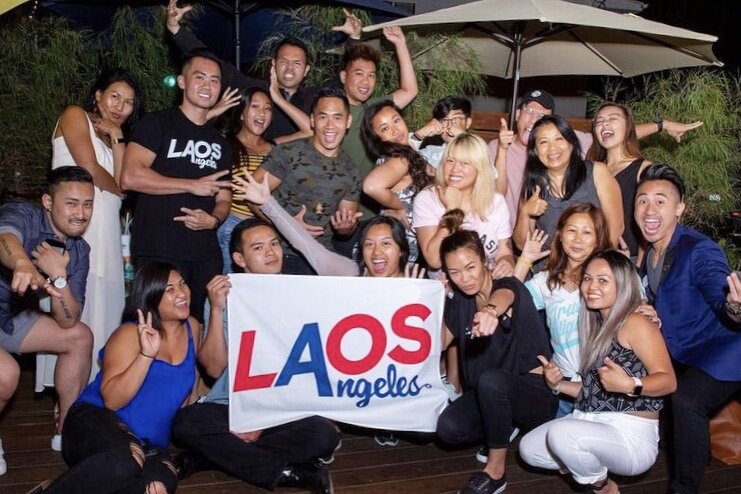 Large group of smiling young people posing at night behind a LAOS Angeles banner
