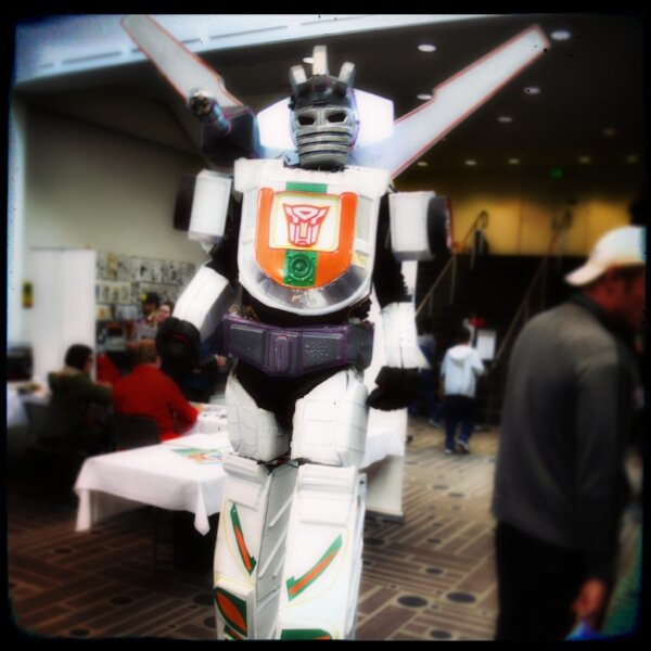 Cosplayer in a homemade white Autobot robot costume walking through a comic con hall.
