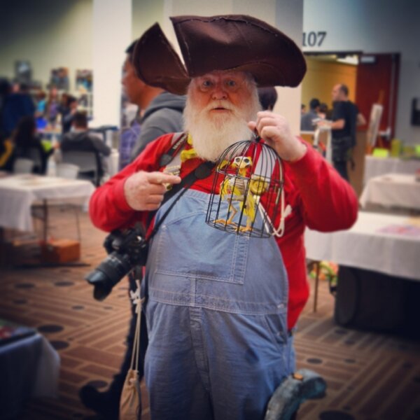 Bearded man in a pirate hat and overalls holding a small birdcage with toy birds at a comic con.