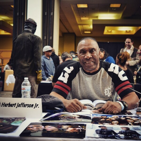 Actor Herbert Jefferson Jr. smiling at his autograph table with signed photos spread out