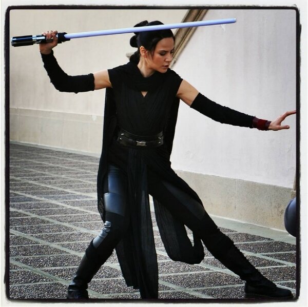 Cosplayer dressed as Rey from Star Wars in a black costume, striking a pose with a blue lightsaber