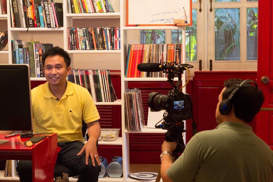 Man in a yellow polo being interviewed on camera in a room lined with bookshelves
