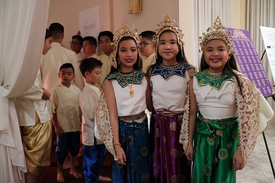 Girls in traditional Lao dance costumes with gold crowns and sinh skirts smile beside boys in traditional outfits