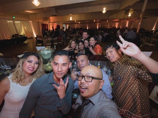 Group selfie of smiling guests making hand gestures in a crowded banquet hall at the Pom Foundation benefit