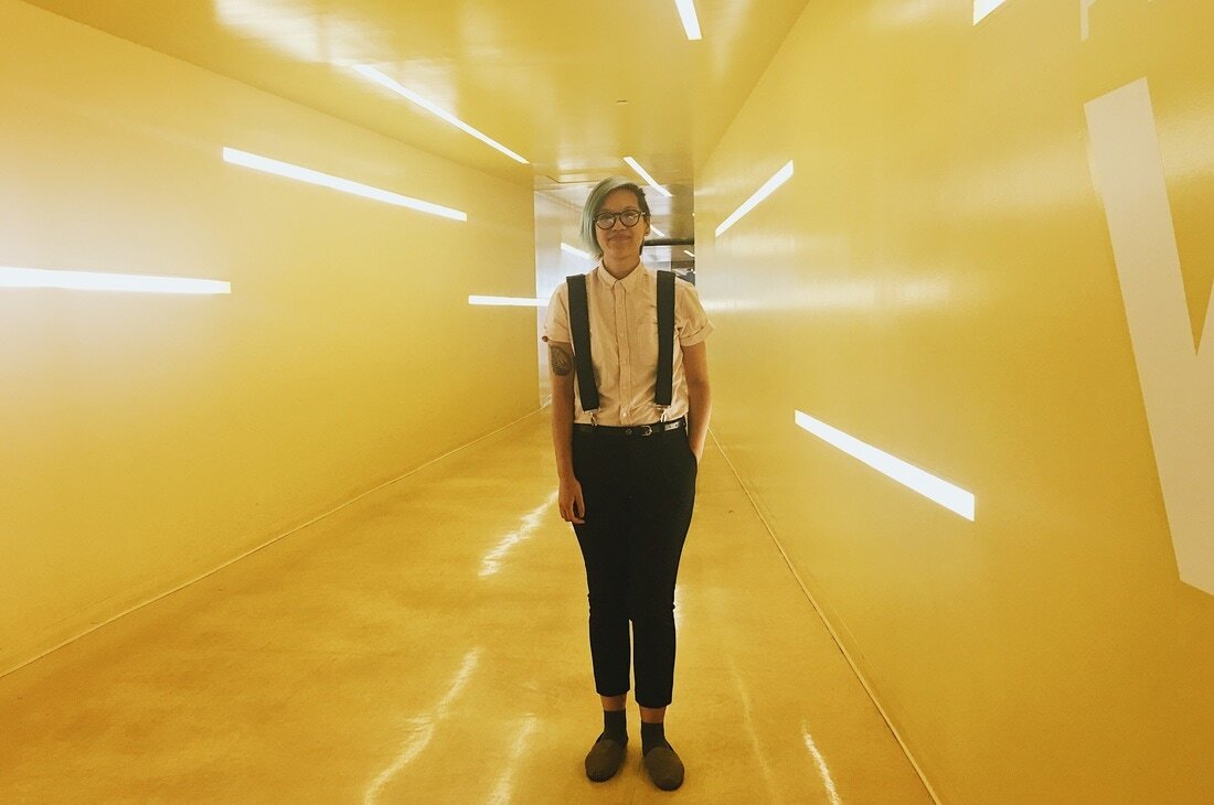 Quyen Nguyen-Le standing in a glowing yellow hallway, wearing suspenders, a white shirt, and glasses