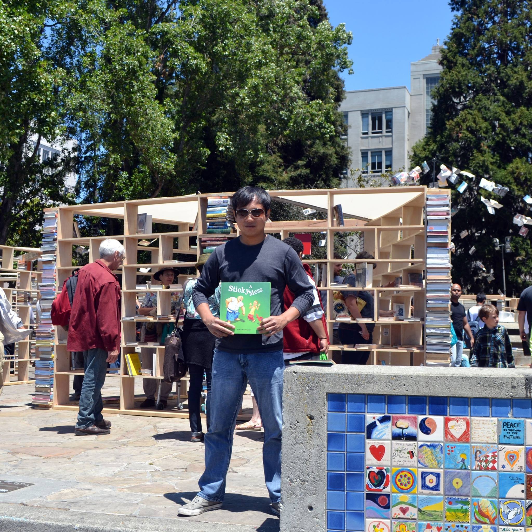 Man in sunglasses holding a copy of A Sticky Mess at the Bay Area Book Festival's outdoor book stalls