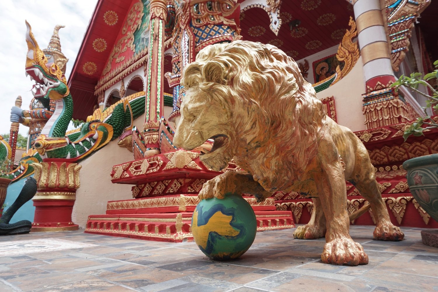 Gold lion statue with its paw on a globe in front of an ornate Lao temple guarded by a green Nak serpent