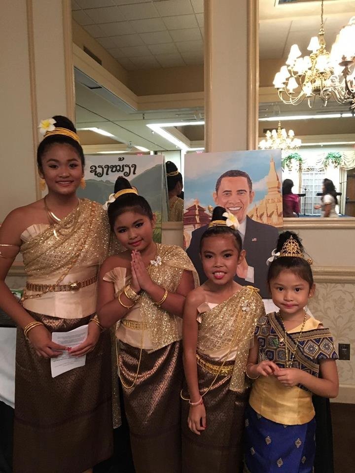 Four girls in traditional Lao dress and sinh skirts at the LACF banquet, one in a nop greeting, beside a painted portrait