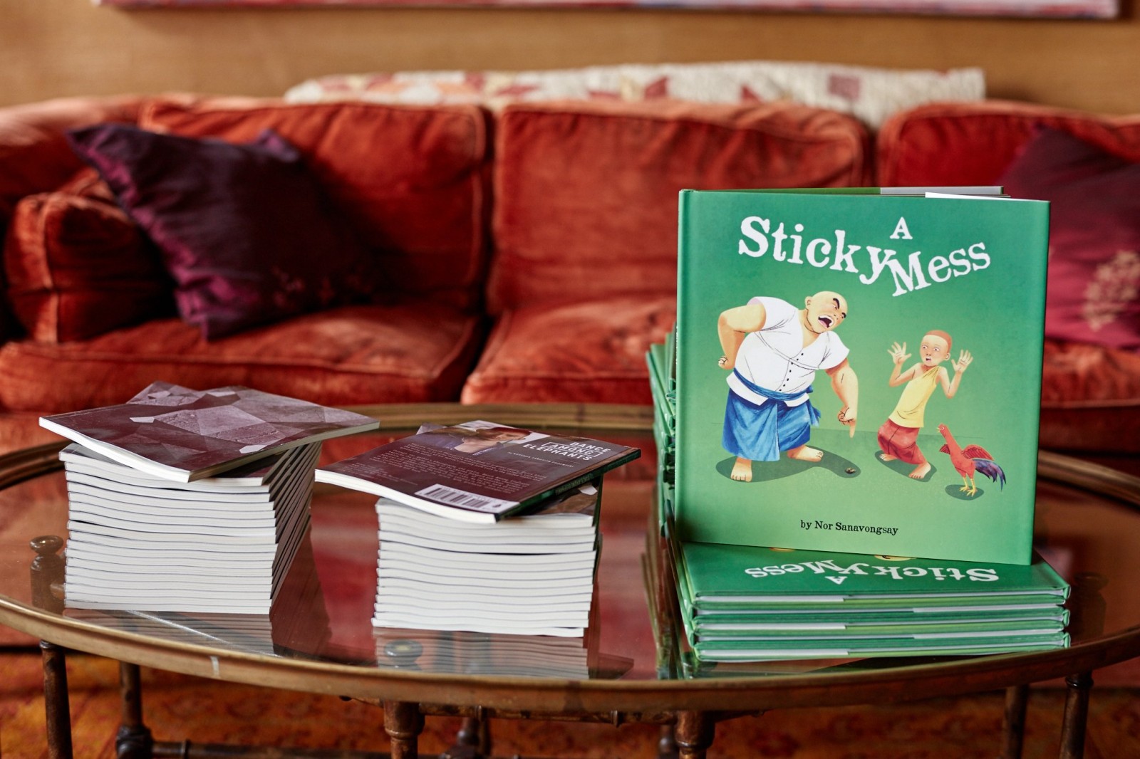 Stacks of A Sticky Mess by Nor Sanavongsay on a glass table, cover showing Xieng Mieng, a boy, and a rooster on green.