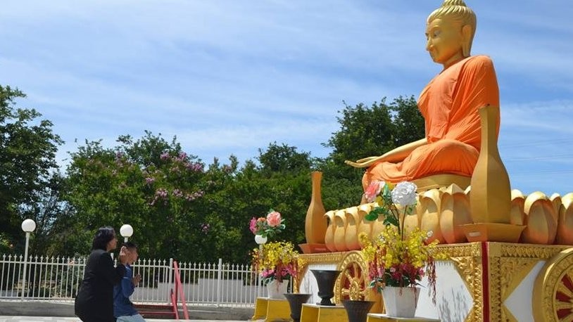 Visitors pray before a large golden seated Buddha statue in orange robes at a temple with flower offerings