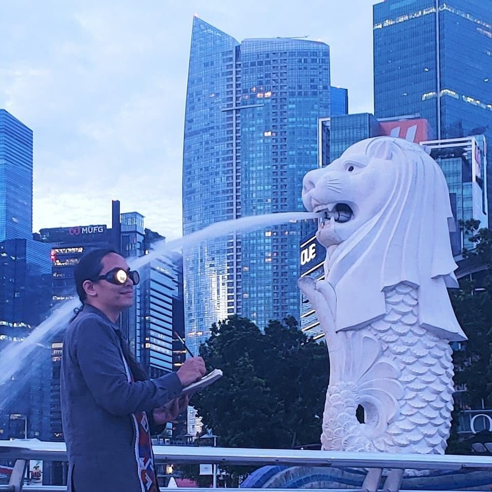Bryan Thao Worra holding a notebook beside the Merlion fountain statue in Singapore, with skyscrapers behind.