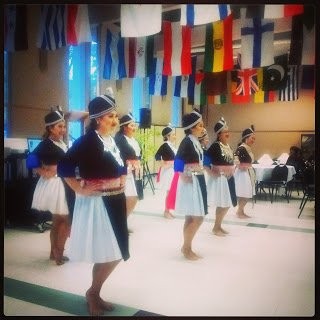 Dancers in black and white traditional costumes with embroidered sashes perform in a hall hung with international flags