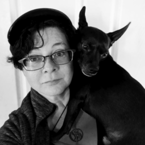 Black and white photo of Angela Yuriko Smith in glasses and a cap, holding a small black dog