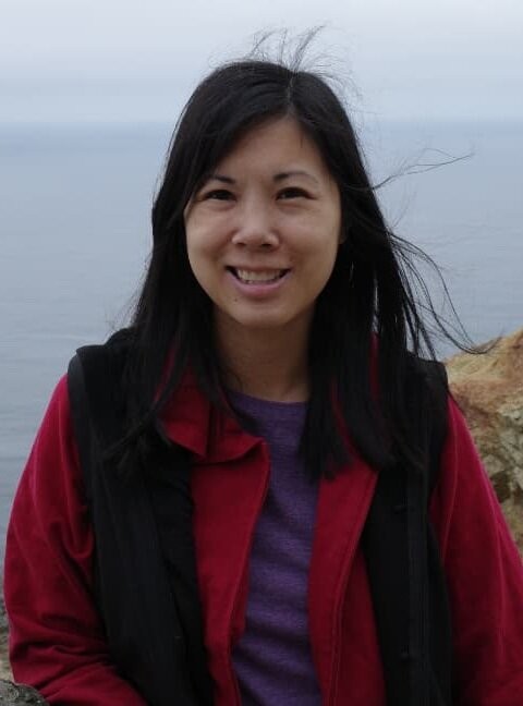 Jenna Le smiling by the ocean in a red jacket, her hair blowing in the wind