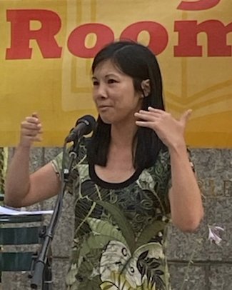 Jenna Le speaking at a microphone outdoors in front of a yellow banner