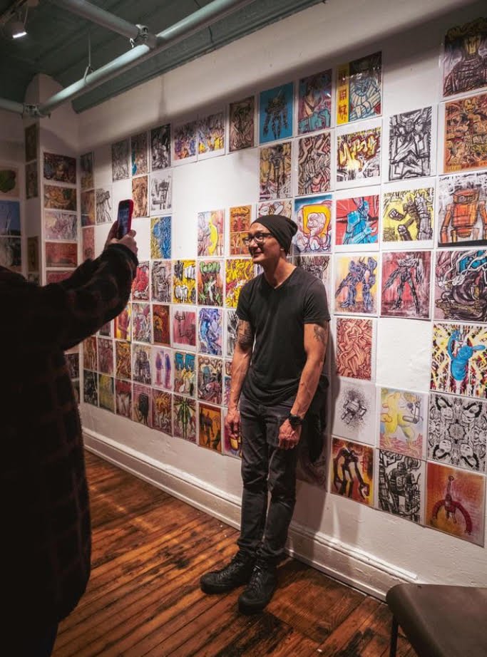Oura Sananikone poses before a gallery wall covered in colorful robot and comic drawings while someone photographs him
