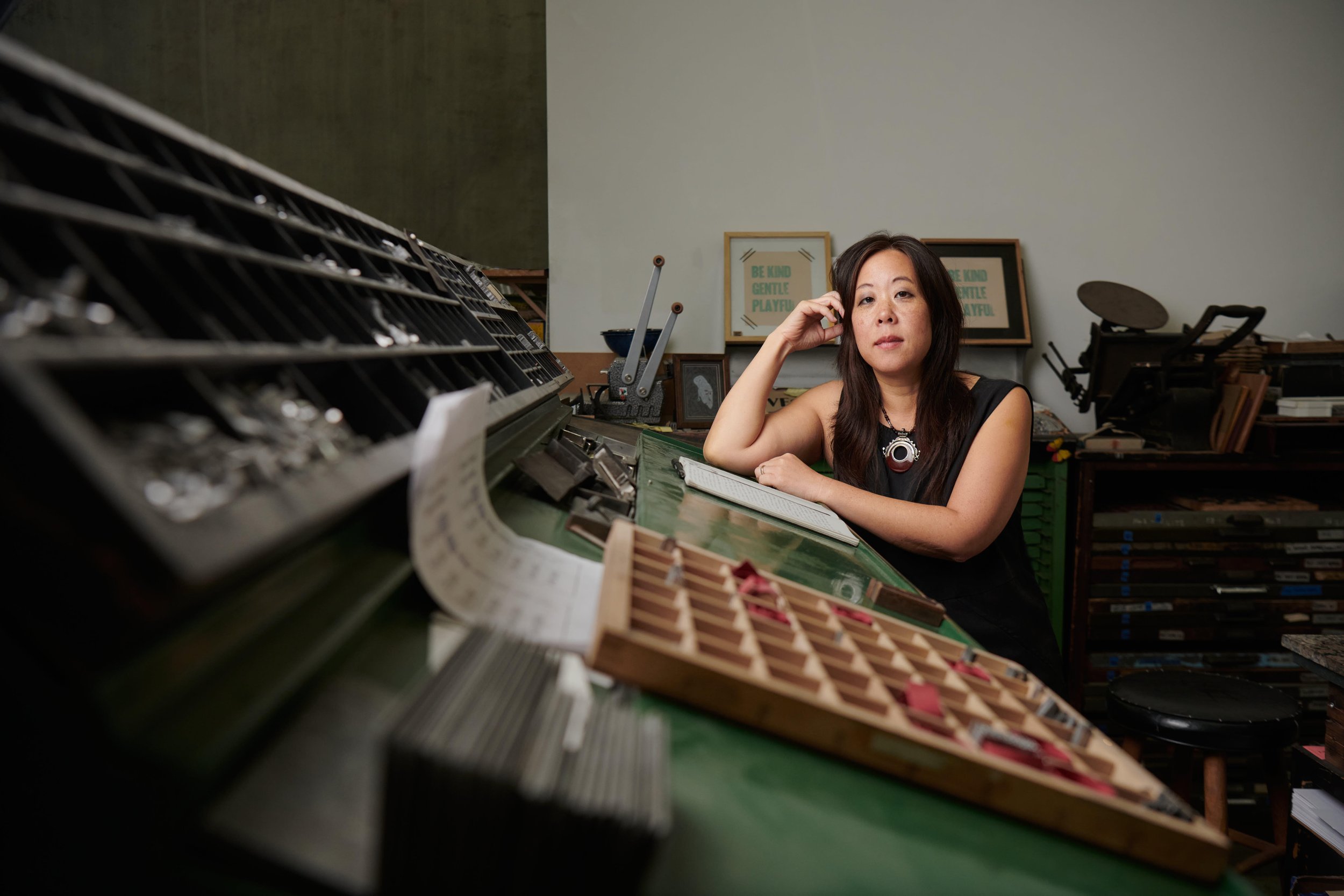 Shin Yu Pai seated at a letterpress print shop table surrounded by trays of metal type