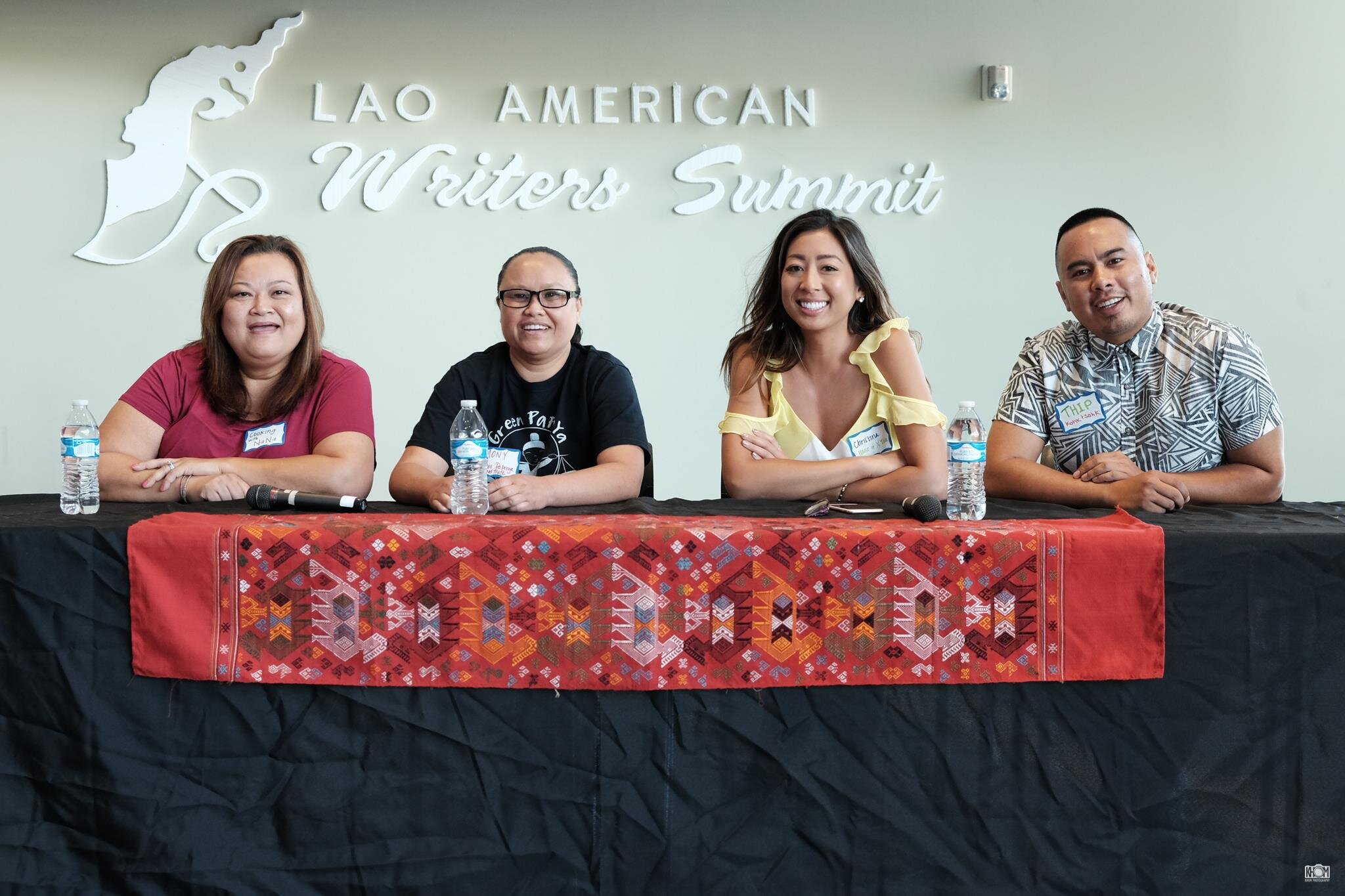Four smiling panelists at a table with a red Lao woven textile runner under the Lao American Writers Summit sign