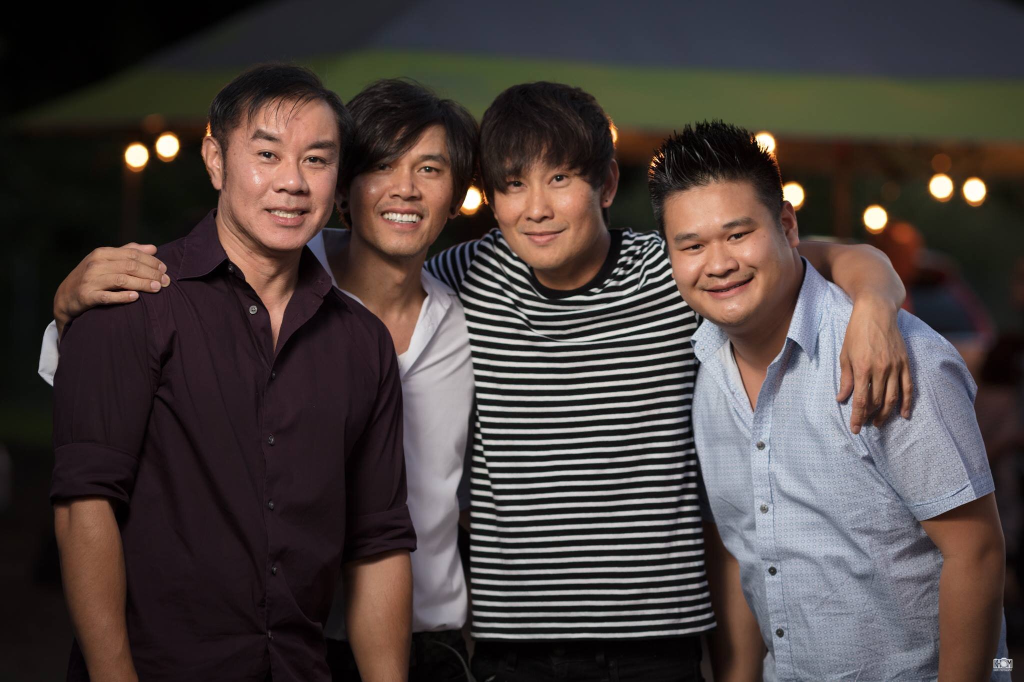 Four men with arms around each other, smiling outdoors at night under string lights