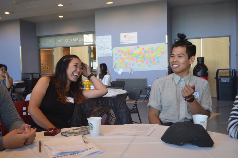 A woman and a man laughing together at a table during a summit session, a wall of colorful sticky notes behind them