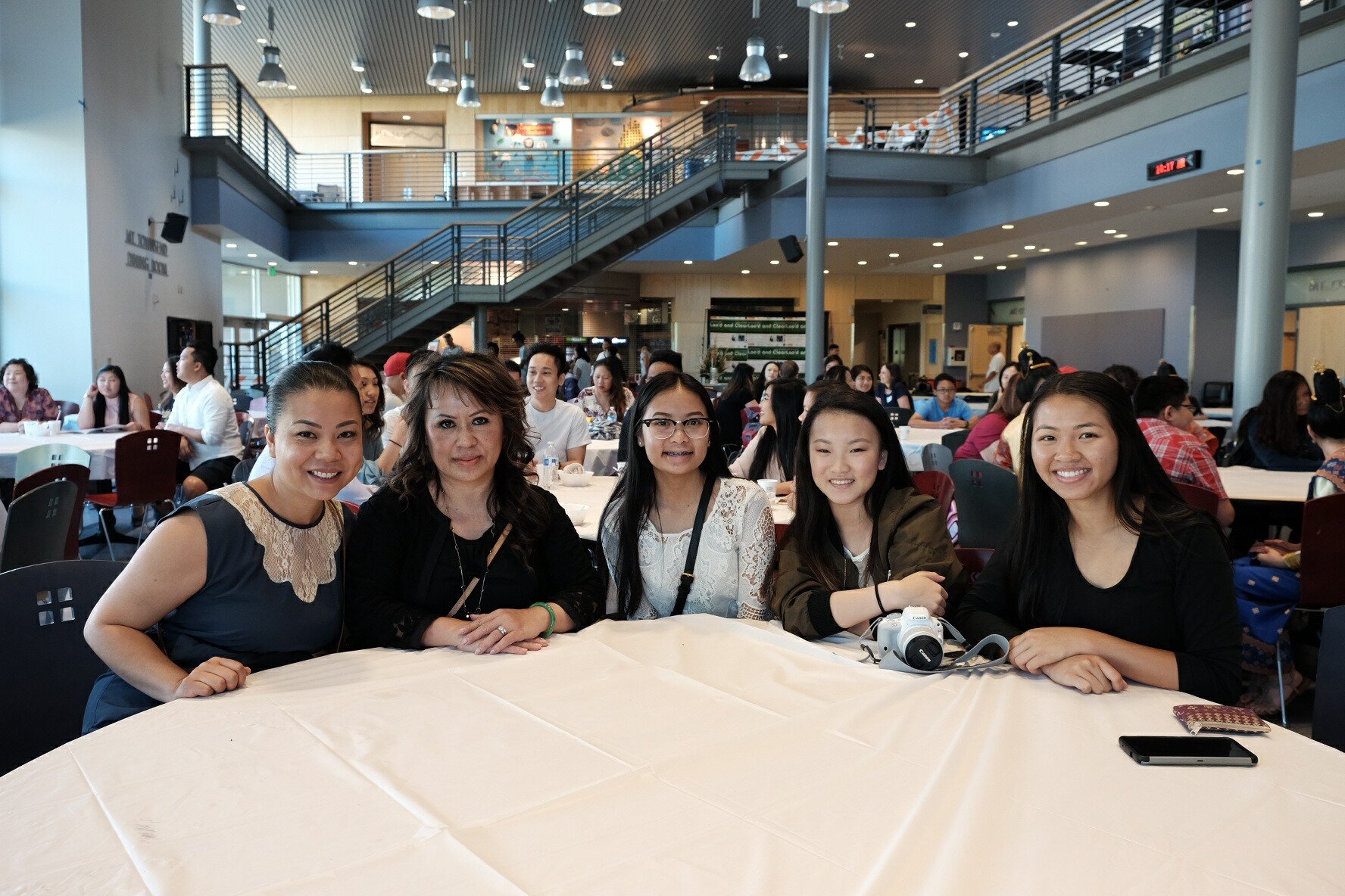 Five women smiling at a round table in a busy campus atrium during the Lao American Writers Summit