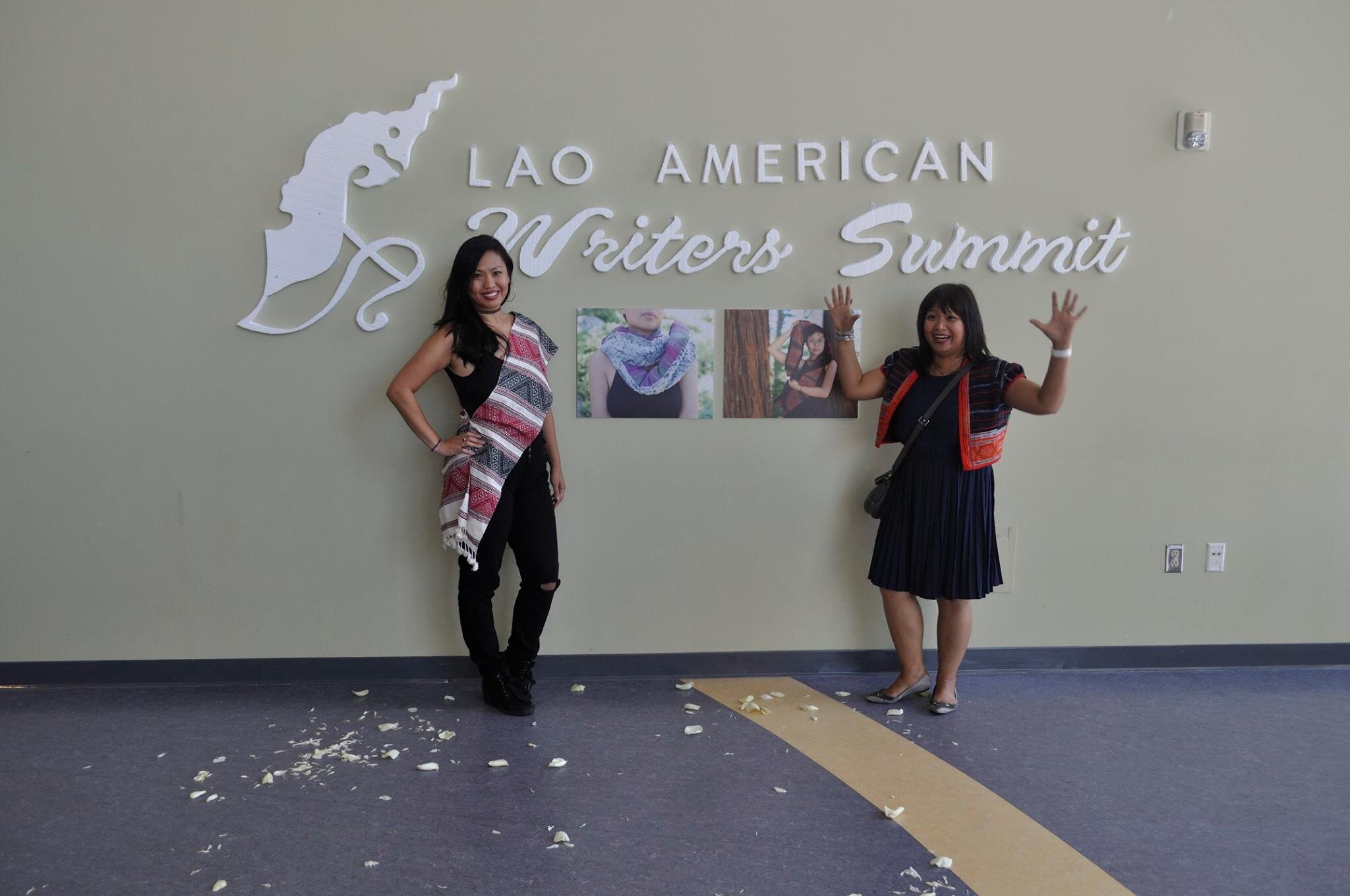 Two women posing on either side of photographs mounted under the Lao American Writers Summit sign, one waving both hands