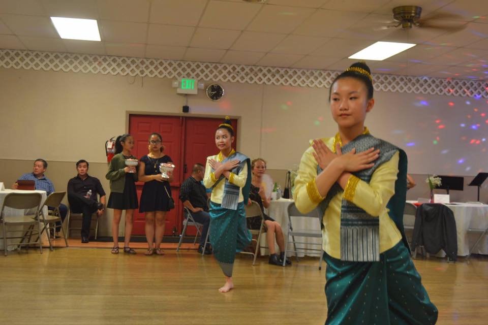 Two young dancers in traditional Lao sinh and sashes performing a Lao dance, hands crossed at the chest, in a community hall
