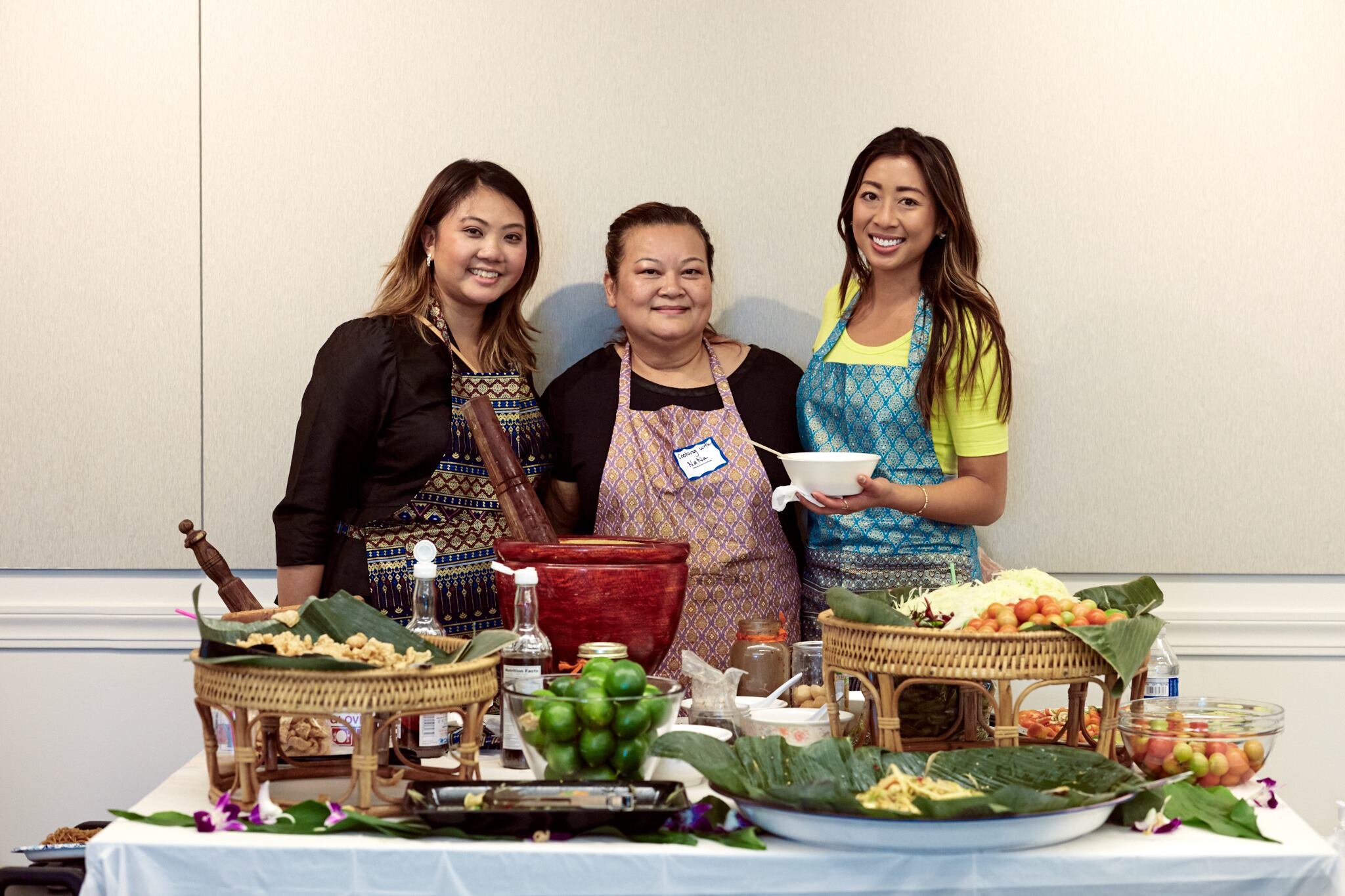 Three women in aprons behind a table of Lao dishes, a clay mortar and pestle, limes, and woven bamboo trays on banana leaves
