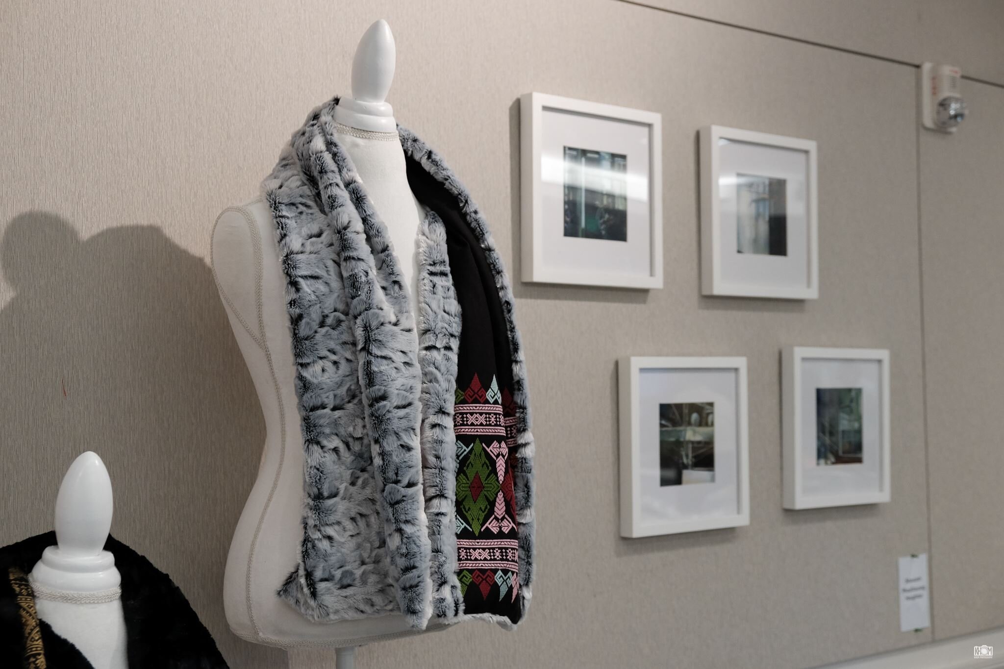 A dress form draped with a fur scarf lined in Lao woven textile, beside framed photographs in an art display