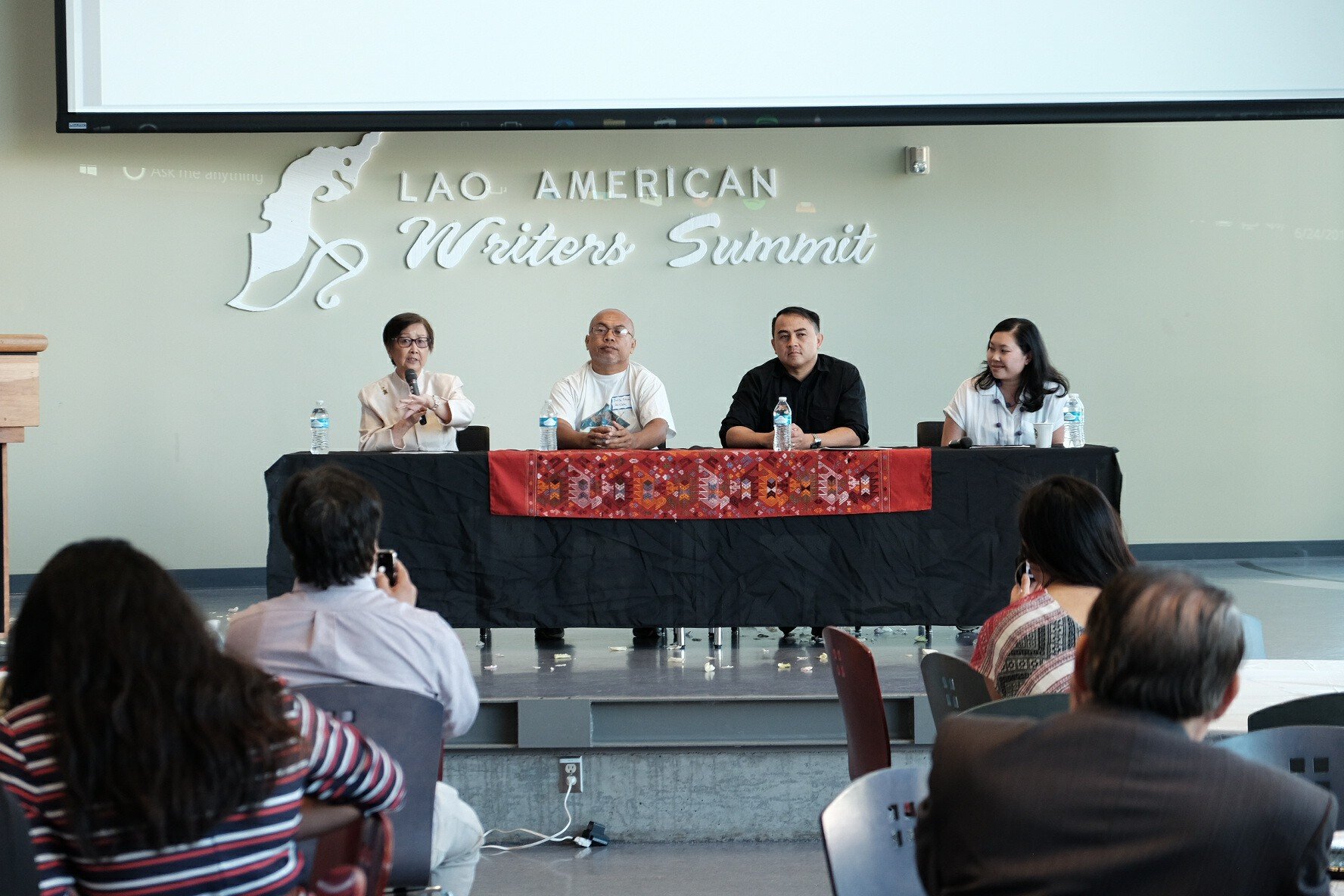 Four panelists seated at a table draped with a Lao textile runner beneath the Lao American Writers Summit sign