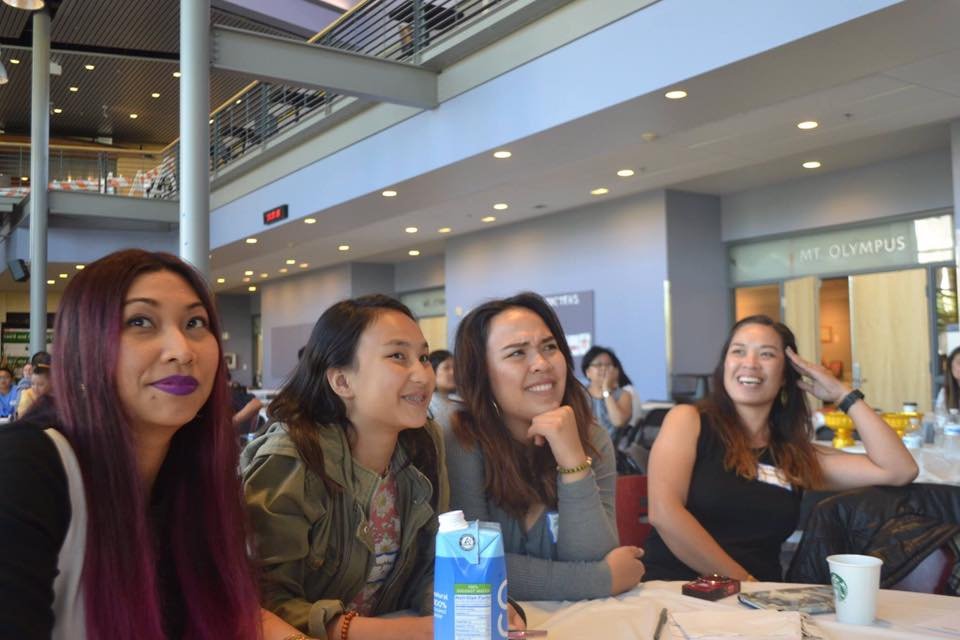 Four women smiling and watching a presentation from their table in the summit atrium