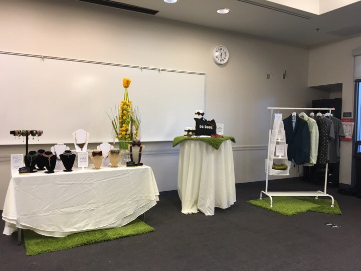Vendor tables set up in a meeting room, with necklaces on display stands, a floral arrangement, and a rack of jackets